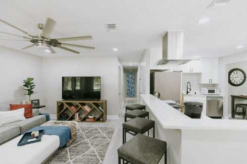 Modern living room and kitchen with a gray sofa, TV, bar stools, and stylish decor in a bright, open space.