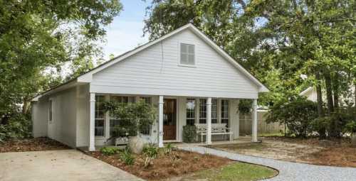 A charming white house with a front porch, surrounded by greenery and a gravel pathway.