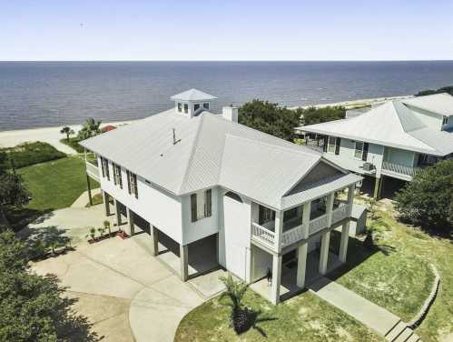 Aerial view of a coastal house with a metal roof, surrounded by greenery and overlooking the ocean.