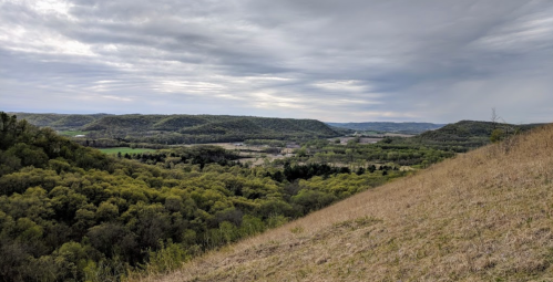 A panoramic view of rolling green hills under a cloudy sky, with a valley and small village in the distance.