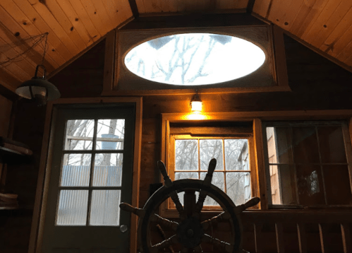 Interior of a wooden cabin with a large oval window, a door, and a ship's wheel in the foreground. Soft lighting.