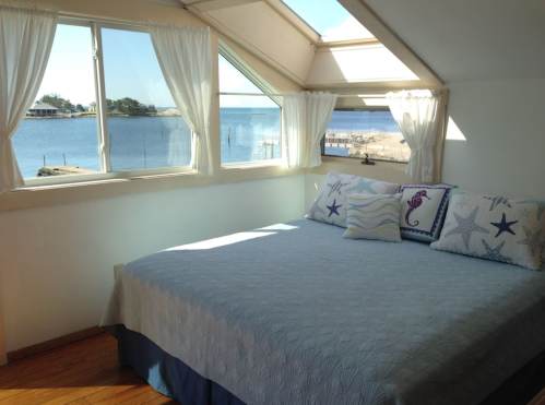 Cozy bedroom with a bed, decorative pillows, and large windows overlooking a scenic waterfront view.