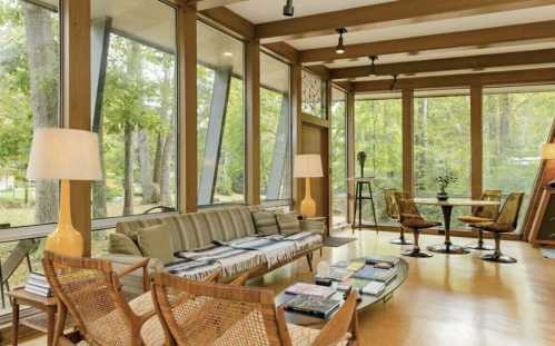 Bright, modern living room with large windows, wooden beams, a sofa, and a dining table surrounded by greenery.