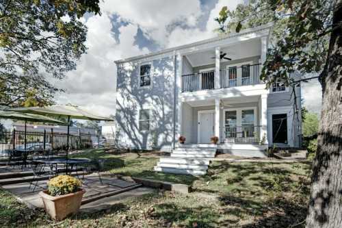 A two-story blue house with a porch, surrounded by trees and a patio area with outdoor seating and flowers.