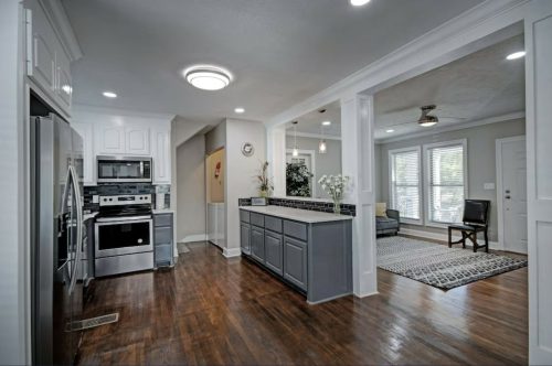 Modern kitchen with stainless steel appliances, gray cabinets, and hardwood floors, leading to a bright living area.