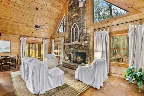 Cozy cabin interior with wooden walls, stone fireplace, and white-draped furniture, surrounded by large windows.