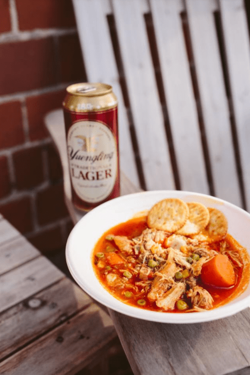 A bowl of chicken soup with vegetables and crackers, next to a can of Yuengling Lager on a wooden table.