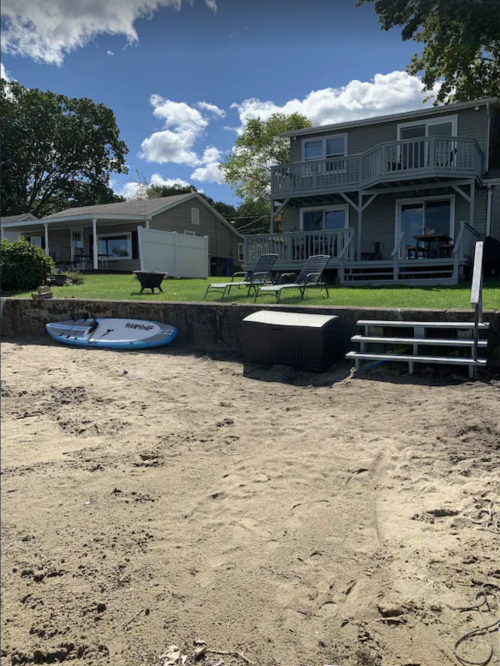 A sandy beach with a surfboard, steps leading to a grassy area, and two houses in the background under a blue sky.