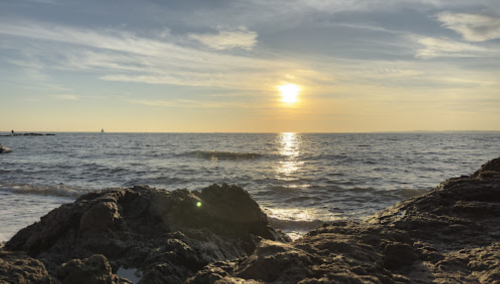 Sunset over the ocean, with gentle waves lapping against rocky shorelines and a warm golden glow in the sky.