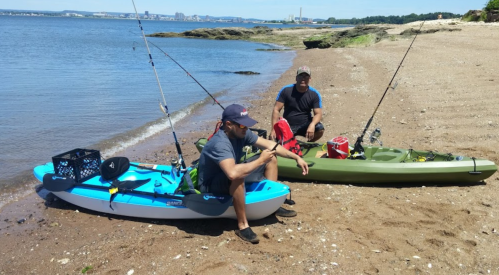 Two men sit on a sandy beach beside their kayaks, equipped with fishing rods, near a calm waterway.