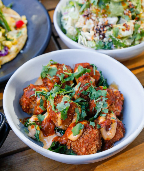 A bowl of crispy meatballs topped with sauce and fresh herbs, served alongside a salad and other dishes.