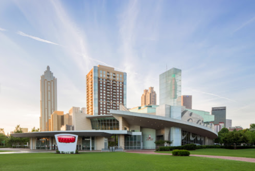 Modern building with a curved roof, surrounded by green grass and city skyscrapers under a clear blue sky.