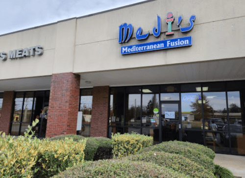 Exterior of Medis Mediterranean Fusion restaurant, featuring a blue sign and large windows, surrounded by greenery.