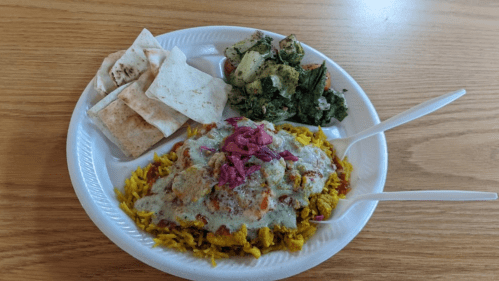A plate of yellow rice topped with sauce, pink onions, and a side of salad and flatbread, served with two spoons.
