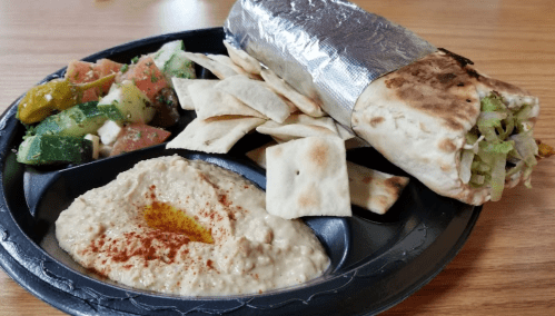 A plate with a wrapped burrito, pita chips, hummus with paprika, and a fresh salad of tomatoes and cucumbers.
