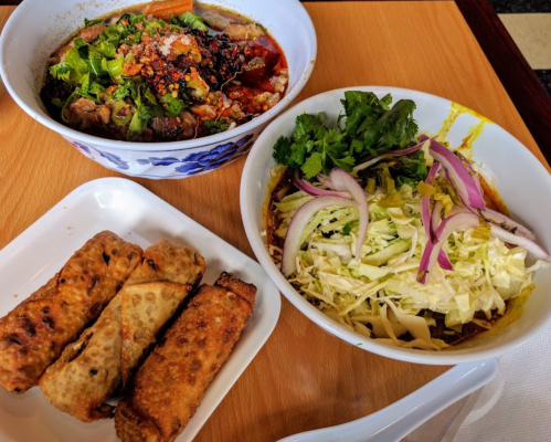 A bowl of spicy soup with herbs, a side salad, and three crispy spring rolls on a wooden table.