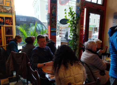 A cozy café scene with people chatting, colorful artwork on the walls, and a view of a bus outside.