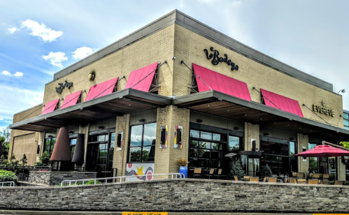 A modern restaurant exterior with pink awnings, large windows, and outdoor seating under umbrellas.