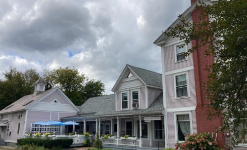 A charming two-story house with a porch, surrounded by trees and a cloudy sky. A smaller building is nearby.