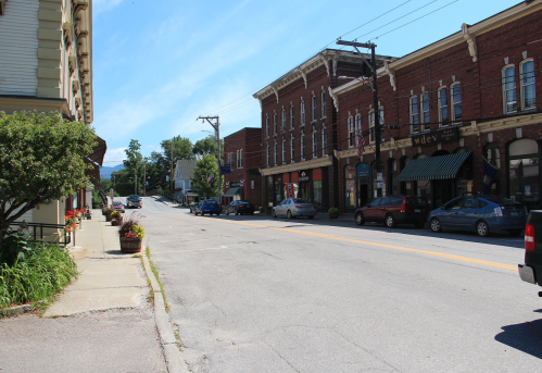 A quiet street lined with brick buildings, shops, and parked cars under a clear blue sky.