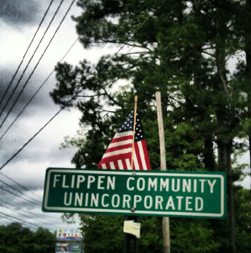 Sign for Flippen Community Unincorporated, with an American flag in the background and trees lining the street.
