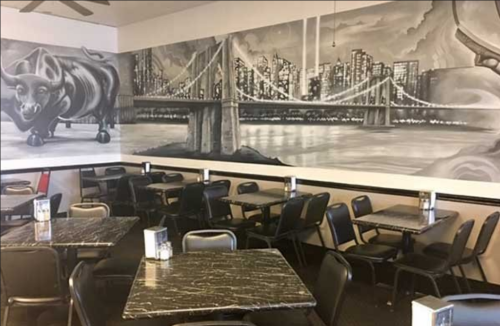 A restaurant interior featuring black and white mural of a bull and a city skyline, with empty tables and chairs.