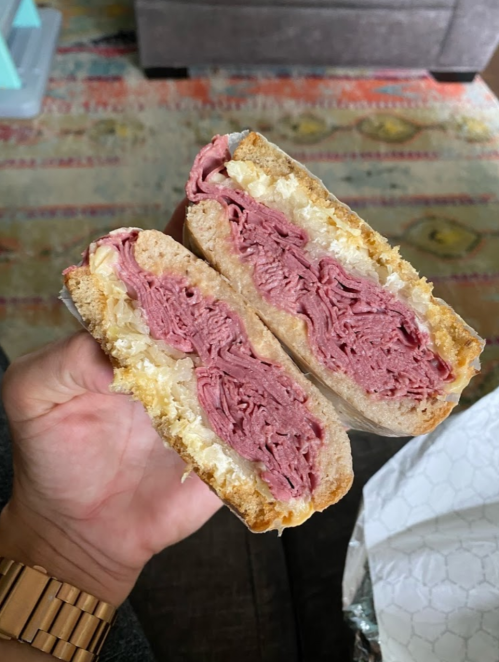 A hand holding a halved sandwich filled with layers of pastrami and sauerkraut on toasted bread.