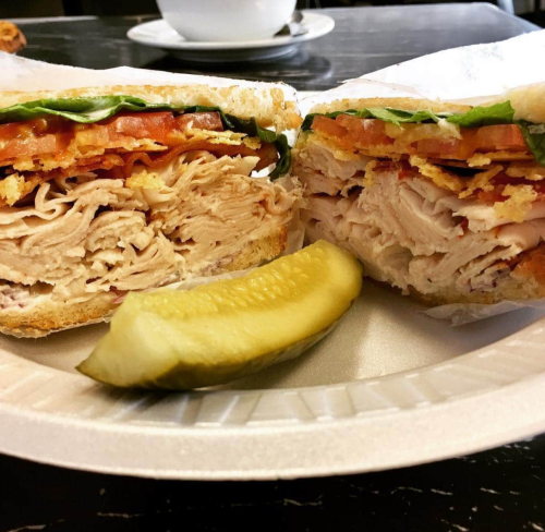 A close-up of a sliced sandwich with turkey, lettuce, and tomato, served with a pickle on a paper plate.