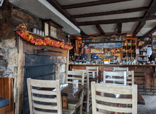 Cozy restaurant interior with wooden chairs, a stone fireplace, and a bar stocked with drinks, decorated for fall.