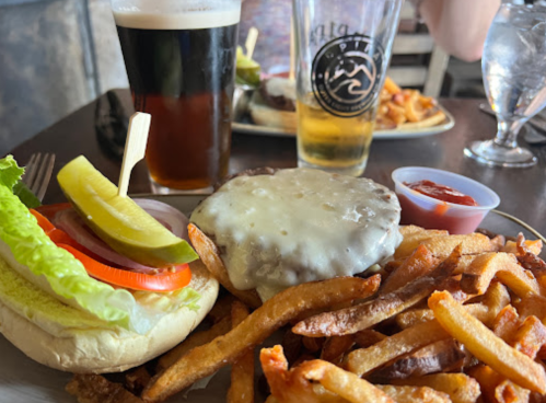 A plate with a cheeseburger topped with lettuce, tomato, and pickles, served with fries and two glasses of beer.