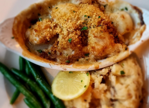 A baked seafood dish topped with breadcrumbs, served with green beans and mashed potatoes, garnished with a lemon slice.