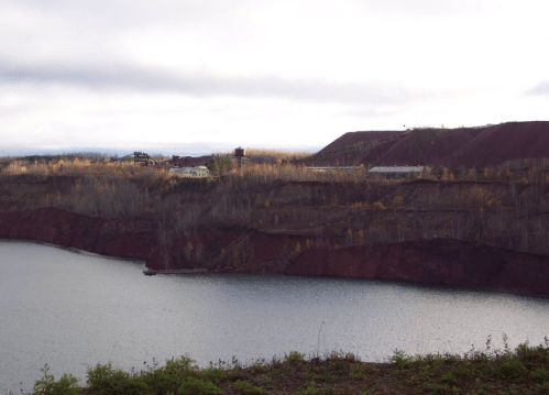 A scenic view of a lake bordered by steep, reddish cliffs and trees, with buildings visible in the background.