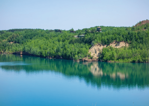 A serene landscape featuring a calm blue lake surrounded by lush green trees and distant structures on a hillside.
