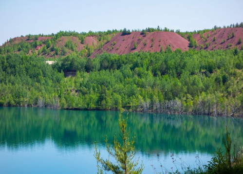 A serene lake surrounded by lush green trees and reddish hills under a clear blue sky.