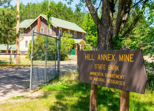Sign for Hill Annex Mine State Park with a building and trees in the background on a sunny day.