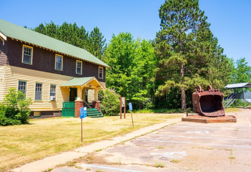 A yellow and brown building beside a large rusted metal object in a grassy parking area with trees in the background.