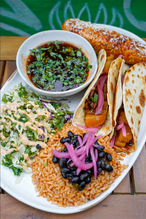 A vibrant plate featuring tacos, rice, black beans, salad, and a bowl of sauce, garnished with herbs.