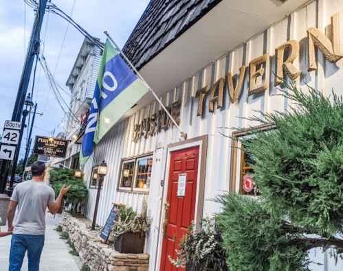 A person walks past a tavern with a flag, surrounded by greenery and street signs in a quaint setting.
