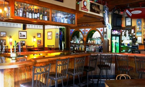 A cozy bar interior featuring a wooden counter, shelves of liquor, and empty stools under warm lighting.