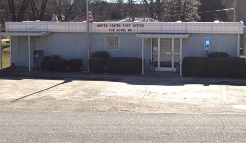 Exterior of the United States Post Office in The Rock, GA, featuring a blue building and a flagpole.