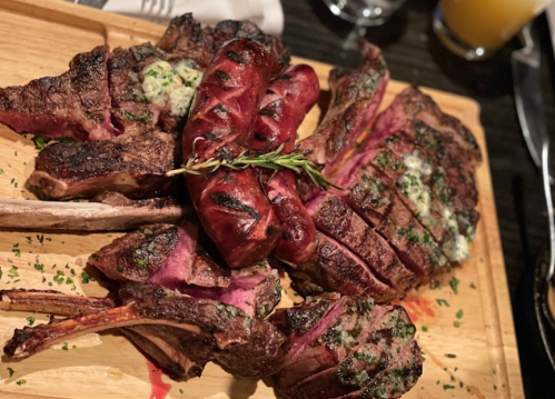 A wooden platter featuring grilled meats, including lamb chops and sausages, garnished with herbs.