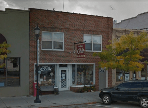 A brick building with a candy shop sign, surrounded by trees and parked cars on a street.
