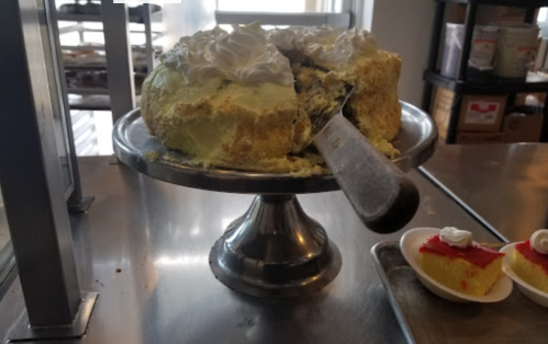 A partially sliced cake with green frosting and whipped cream on a silver cake stand, next to a slice of red dessert.