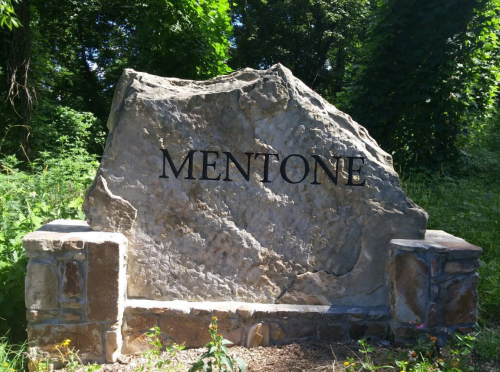 A large stone monument with the word "MENTONE" engraved on it, surrounded by greenery.