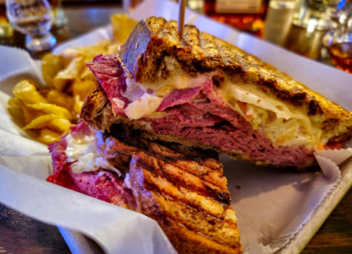 A close-up of a grilled sandwich filled with layers of meat and cheese, served with crispy potato chips on the side.