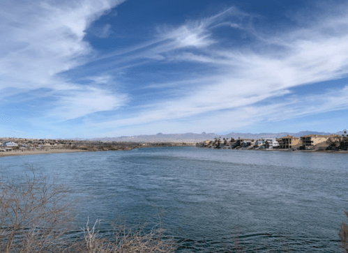 A serene river flows under a blue sky with wispy clouds, bordered by homes and distant mountains.