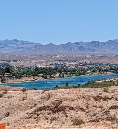 A scenic view of a river winding through arid land, with mountains in the background under a clear blue sky.