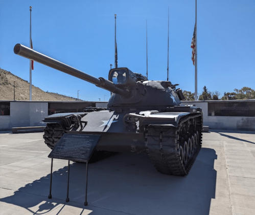 A military tank displayed outdoors, with flags in the background and a plaque in front on a clear day.