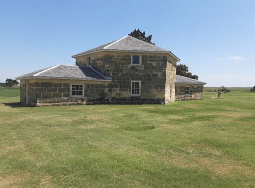 A stone building with a gray roof, surrounded by green grass under a clear blue sky.