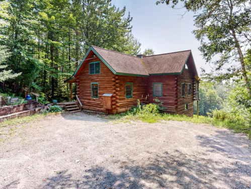 A rustic log cabin surrounded by trees, with a gravel driveway and a peaceful, natural setting.